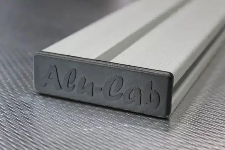 Close-up of a silver rectangular metal bar with a rounded end and cursive engraving on the end face on a textured surface.