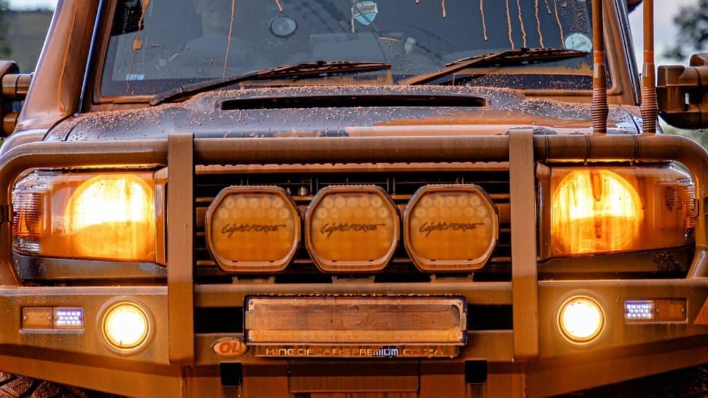 Front view of a muddy orange off-road truck with a sturdy grille guard and three circular bumper lights