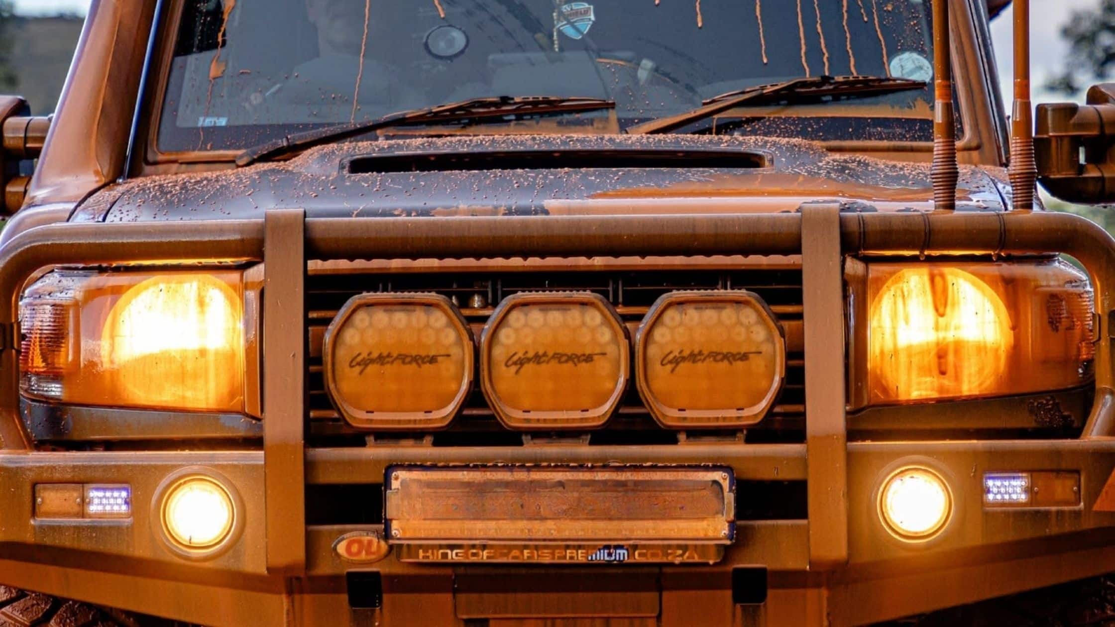 Front view of a muddy orange off-road truck with a sturdy grille guard and three circular bumper lights