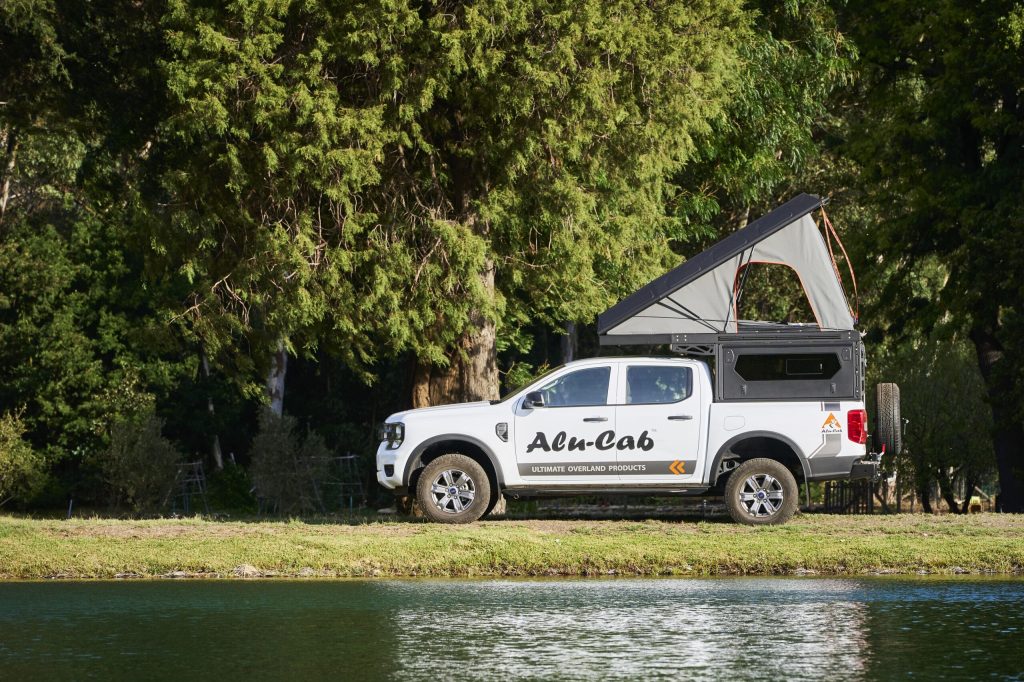 White pickup truck with a rooftop tent parked on a grassy lakeshore, dense trees in the background.